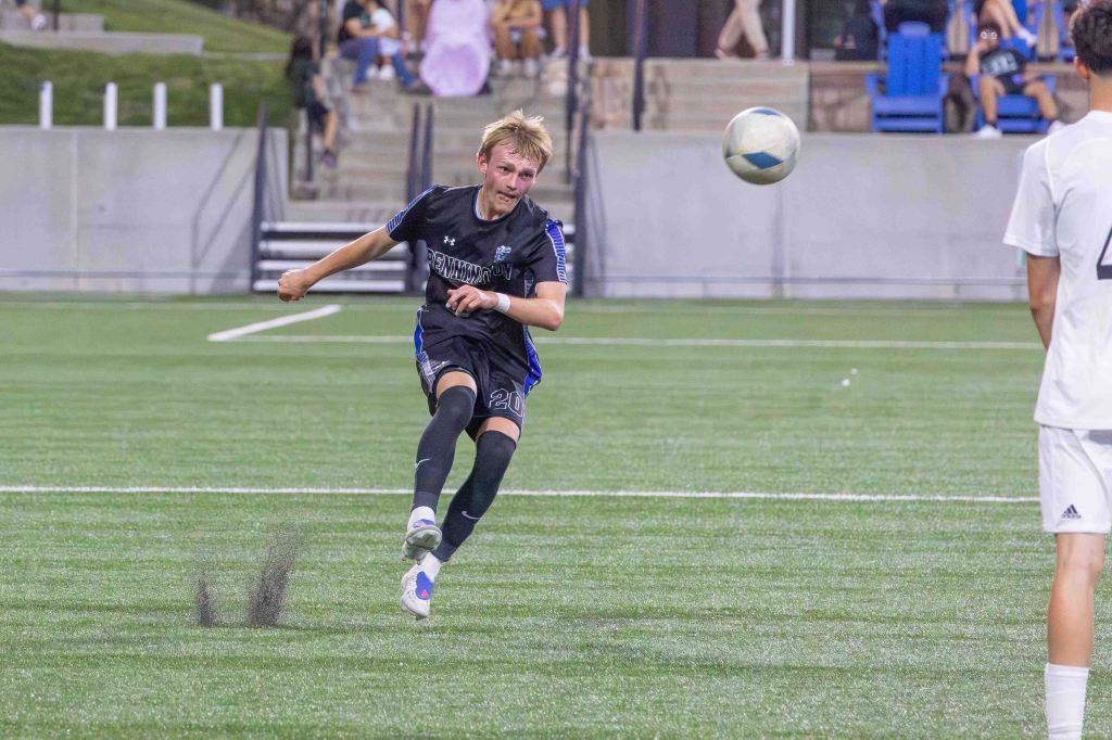 A young soccer player in a black uniform kicks a soccer ball on the field while another player in a white uniform watches.
