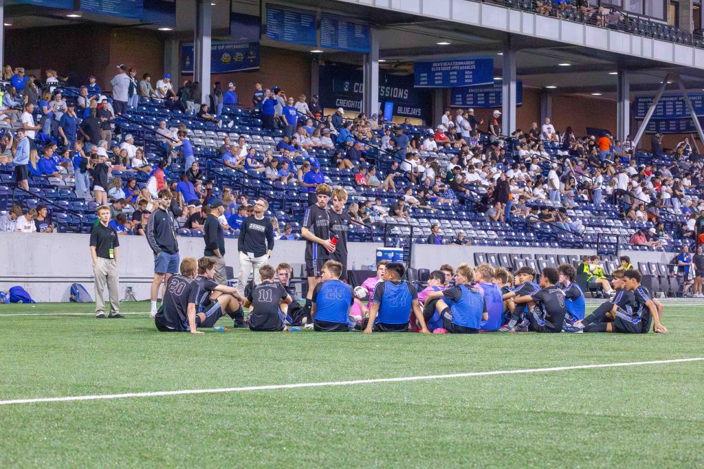 A youth soccer team sits in a circle on the field, listening to their coach who stands among them. In the background, a large crowd fills the stadium stands, with many fans dressed in blue and white.