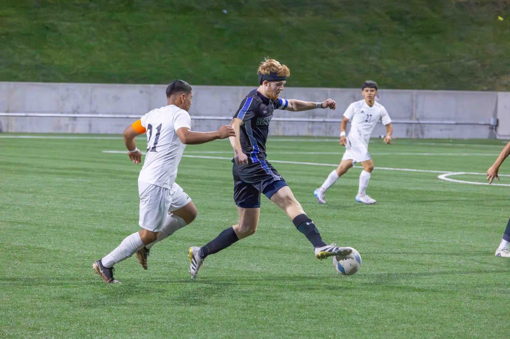 A soccer player in a black jersey kicks the ball while being closely pursued by an opponent in a white jersey on a green field.