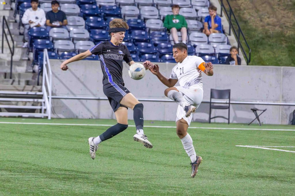 Two young soccer players competing for the ball on a grassy field during a game, with spectators seated in the background.