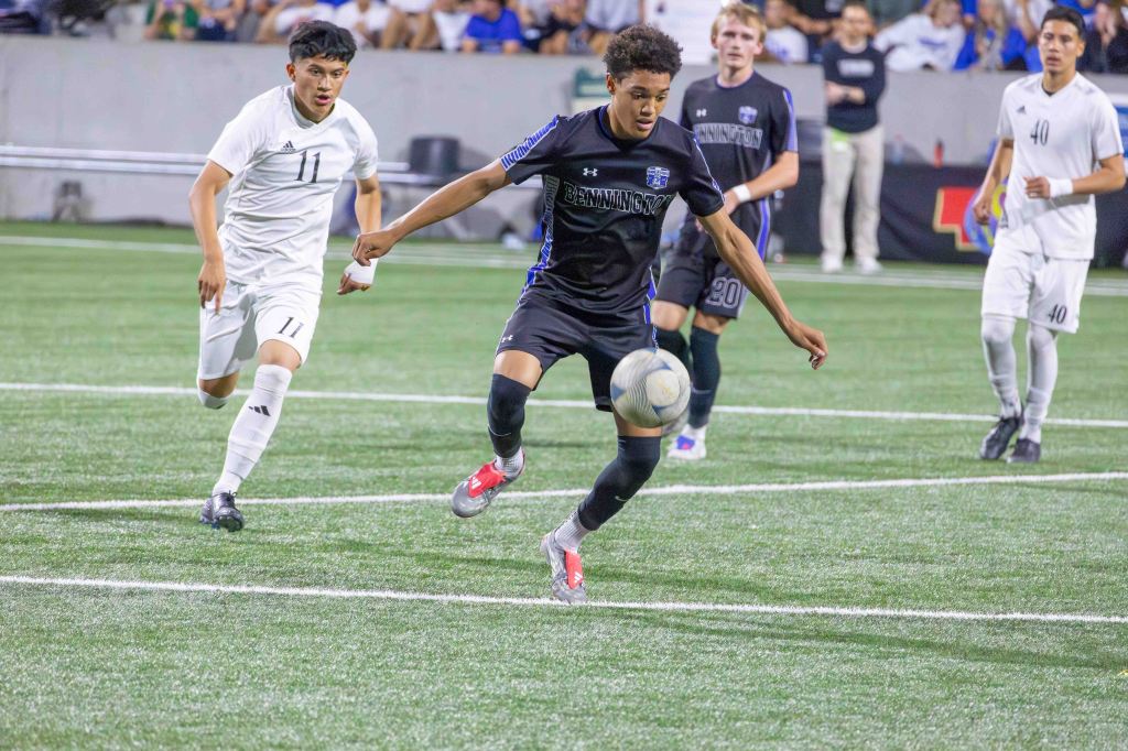 A soccer player in a black uniform dribbles the ball on a green field, while an opposing player in a white uniform closely follows behind.