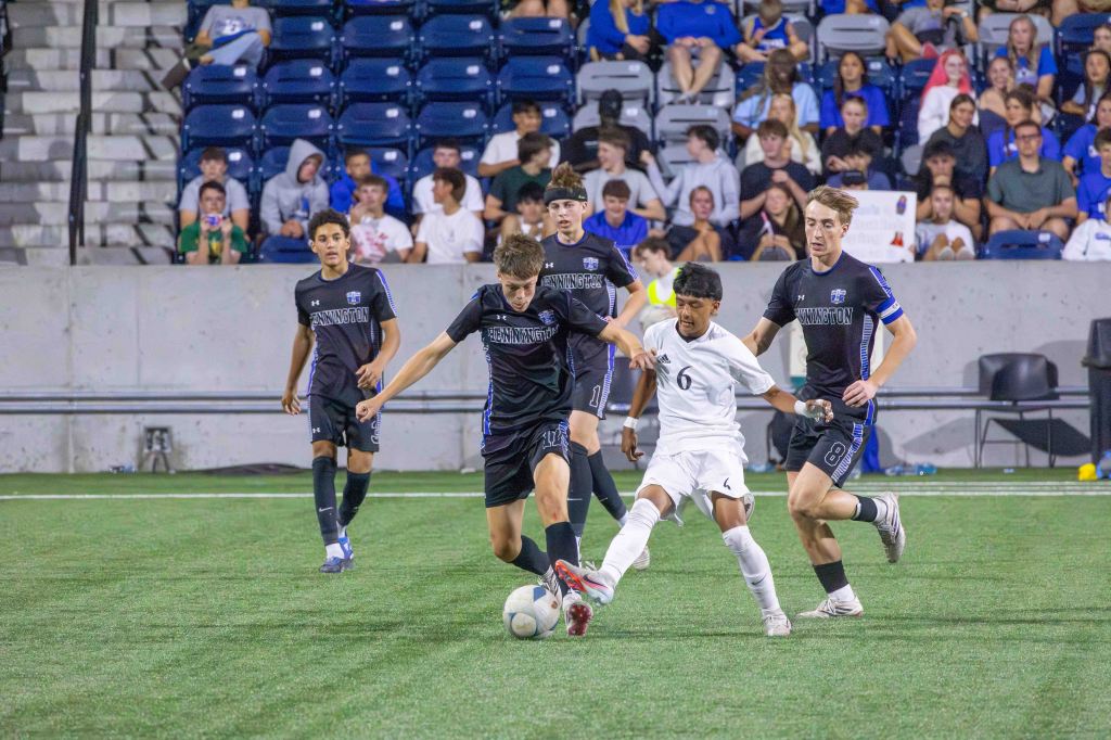 A soccer match in progress with players wearing black and white uniforms. One player in white is trying to gain control of the ball as he is challenged by two players in black. A crowd is visible in the background, cheering and watching the game.