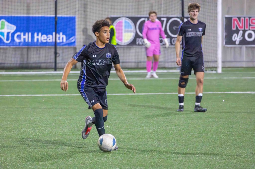 A young soccer player in black and blue uniform dribbles a soccer ball on a turf field, with teammates and a goalkeeper in the background.