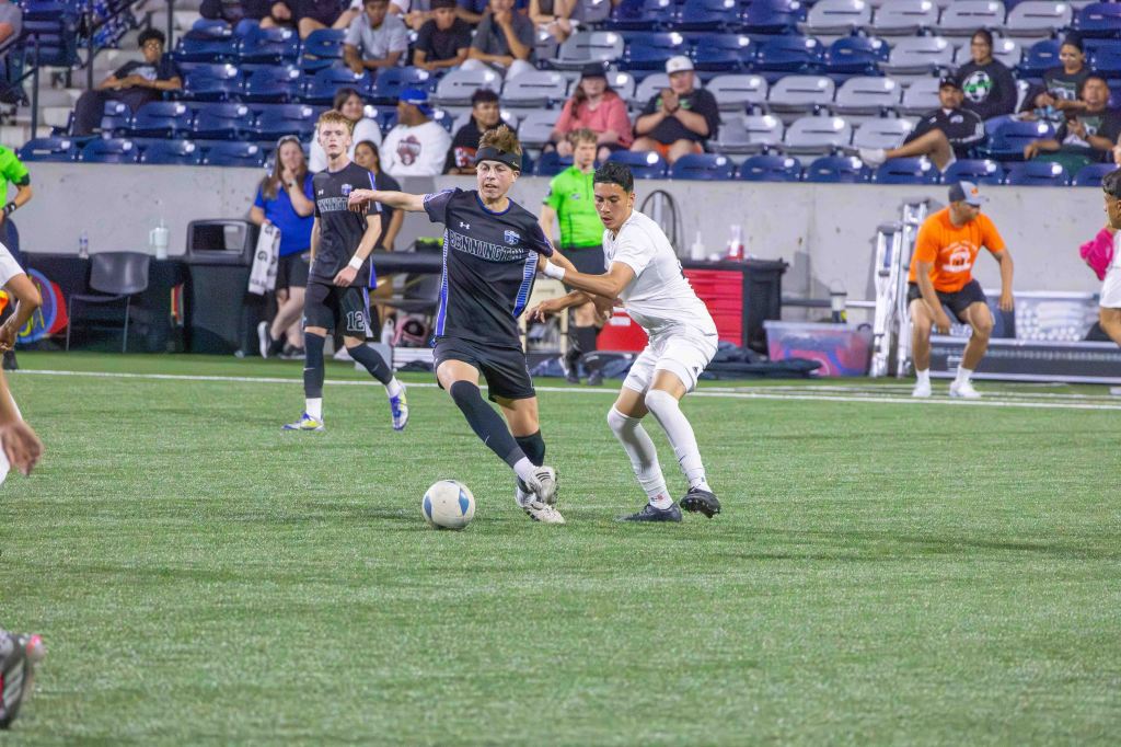A soccer match in progress featuring two players competing for the ball on a synthetic field. One player in black is attempting to maintain possession while another in white is challenging for it. Spectators can be seen in the background.