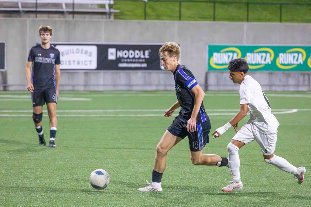A soccer match in progress with two players in action; one in a black jersey with the number 8 and a captain's armband chasing a soccer ball, while a player in a white jersey tries to regain possession. Another player in a black jersey is observing the play in the background.