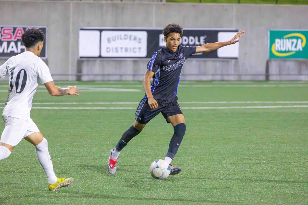 A male soccer player in a black jersey dribbles the ball on a field while another player in a white jersey approaches from the left.