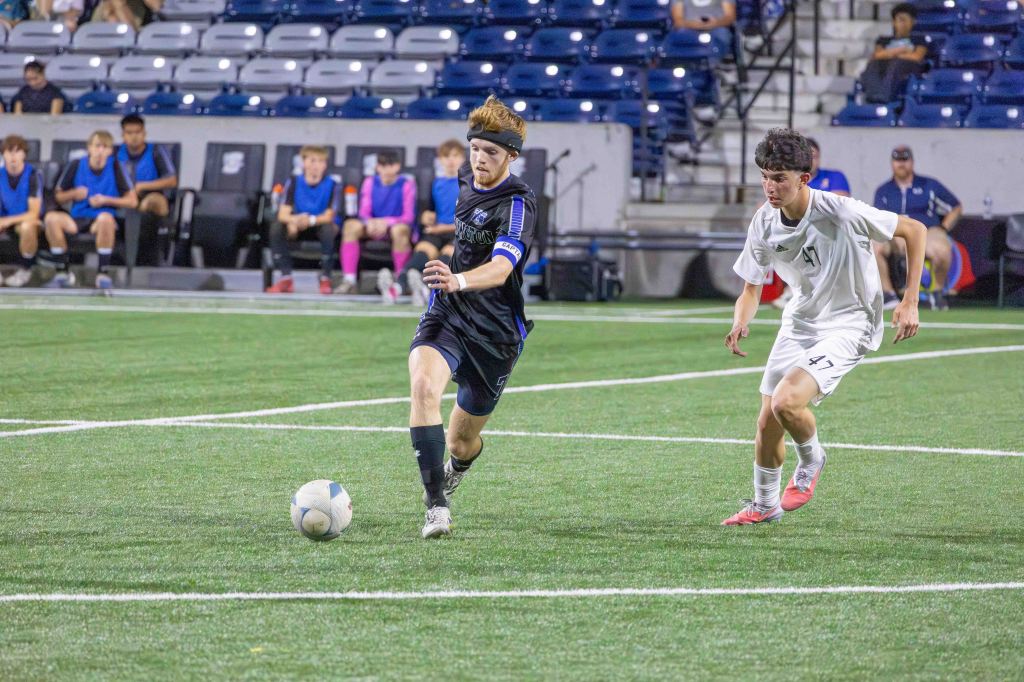 A soccer player in a blue uniform dribbling the ball on a green field, with a player in a white uniform sprinting to defend.