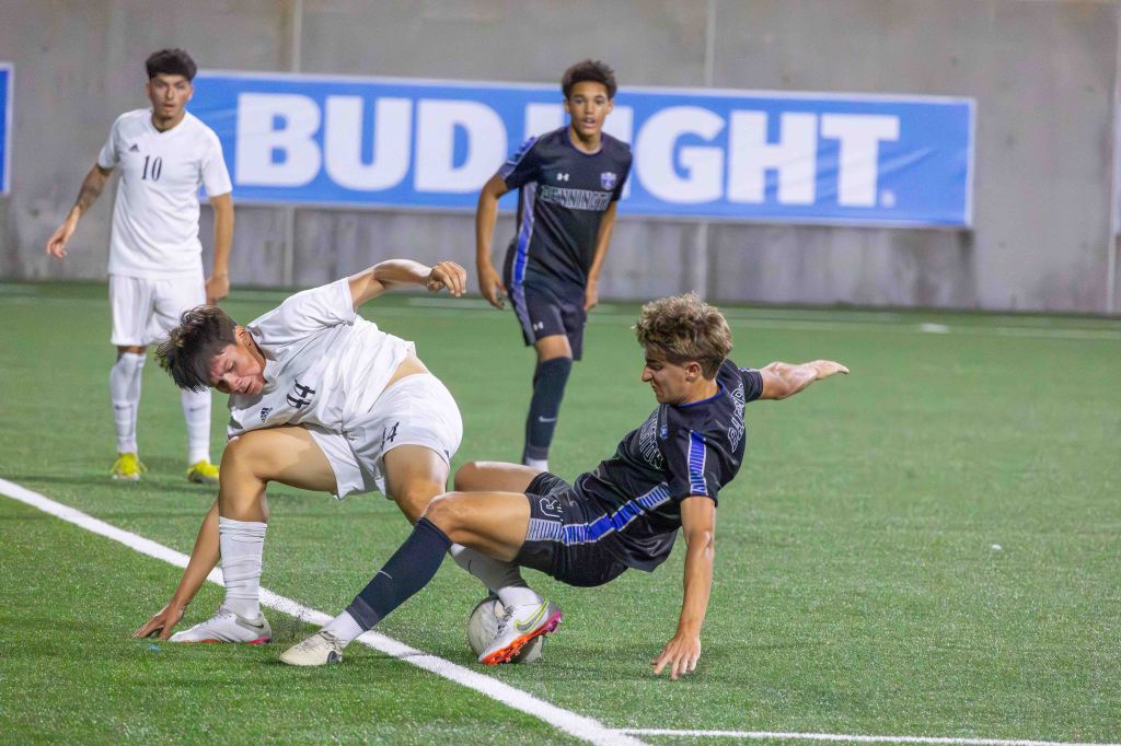 Two youth soccer players competing for the ball, one player in a white jersey is falling to the ground while the other in a black jersey is attempting to tackle.