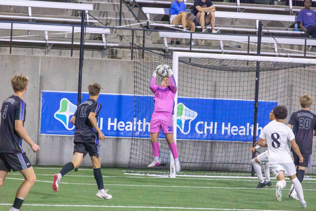 A goalkeeper in a pink uniform catches a soccer ball during a game, with players from both teams in the background and a sponsorship banner visible.