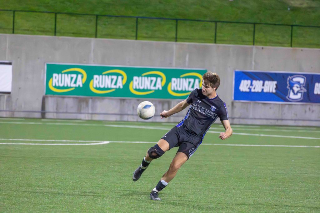 A male soccer player in a black jersey with blue accents kicks a soccer ball during a game on a green field, with advertisements visible in the background.
