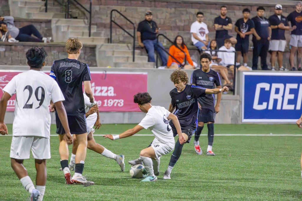 A high school soccer game scene showing players in action on the field. A player in a white uniform is attempting to take the ball from an opponent in a dark uniform. Spectators are visible in the background.
