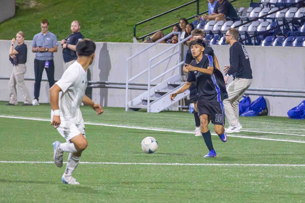A soccer player in a black jersey with the number 10 is about to kick a soccer ball on a field, while another player in a white jersey approaches from the left. In the background, spectators and coaches watch the game.
