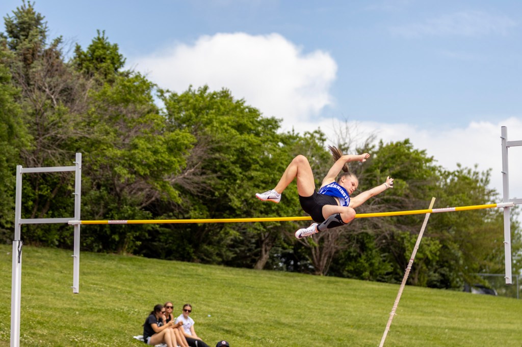 A female athlete performing a high jump over a yellow bar on a grassy field, with spectators sitting in the background.