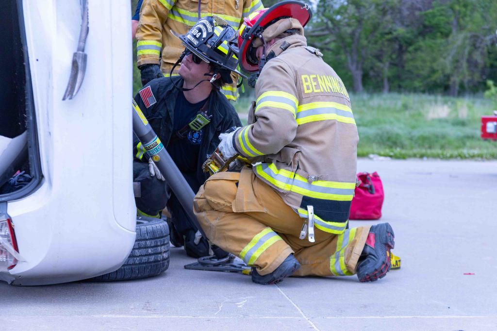 Two firefighters working together near a flipped white vehicle during a rescue operation. One firefighter is holding a rescue tool while the other is kneeling and assisting.