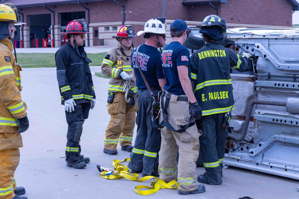 A group of firefighters in uniform is gathered around a flipped-over vehicle, discussing rescue techniques. The scene takes place outside a fire station, with fire equipment visible.