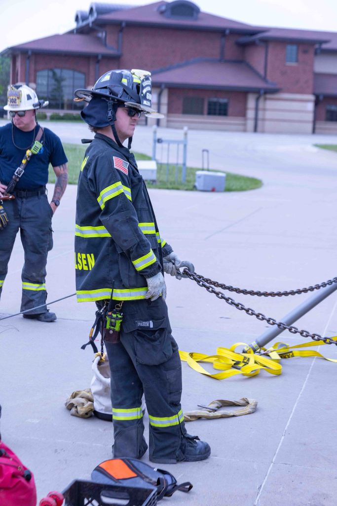 Firefighter standing on a pavement holding a chain, wearing protective gear, with another firefighter in the background.