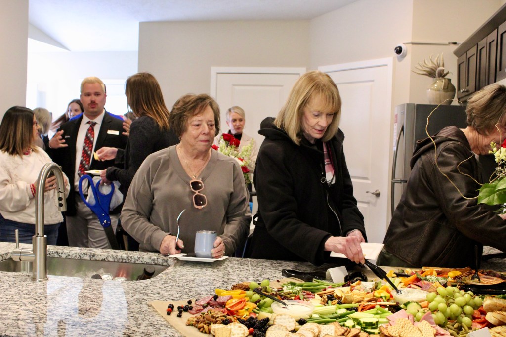 A group of people socializing at a gathering, with two women preparing food from a large charcuterie spread on a kitchen counter. The background includes other attendees engaged in conversation.