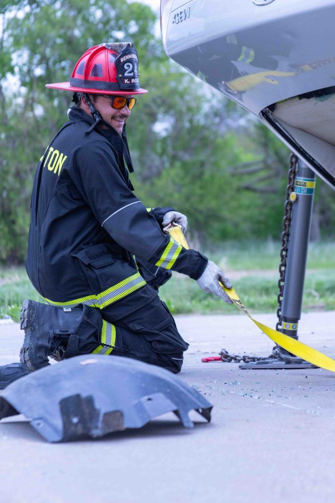 A firefighter in protective gear kneels on the ground, working with a yellow strap and chain near a vehicle. Detritus from the scene, including a piece of plastic, is visible on the ground.