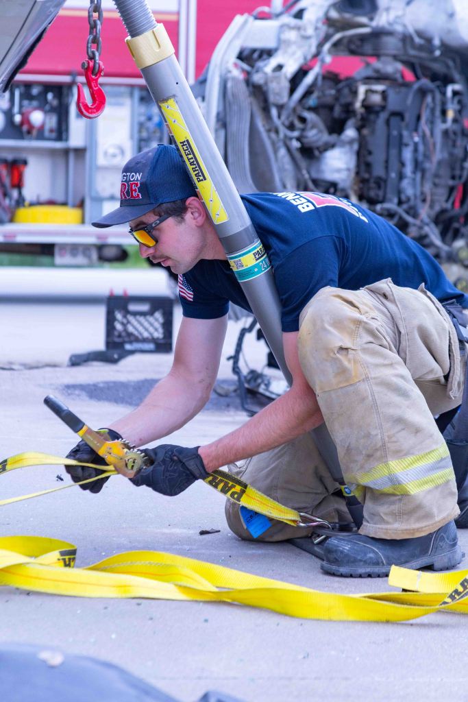 A firefighter crouching down on a pavement, working with yellow straps and a tool, with a vehicle in the background.