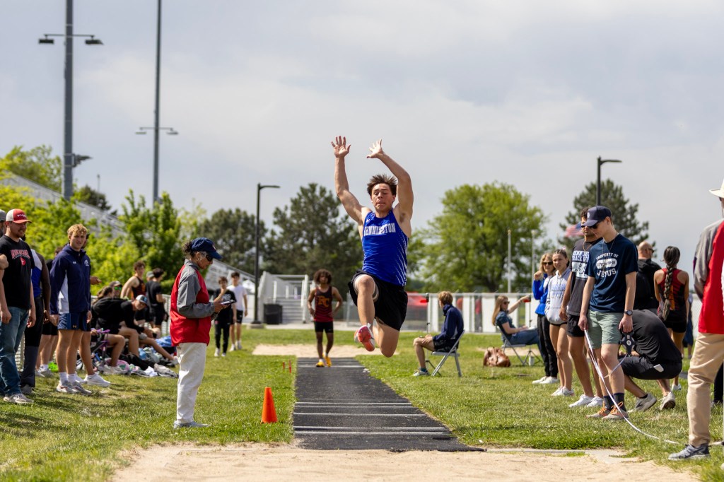 An athlete in a blue jersey is performing a long jump at a track and field event, with spectators watching from the sidelines.