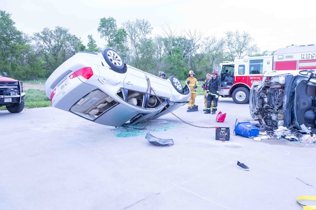 A scene of a car accident featuring an overturned vehicle and a fire truck. Emergency responders, including firefighters, are present at the scene, assessing the situation.