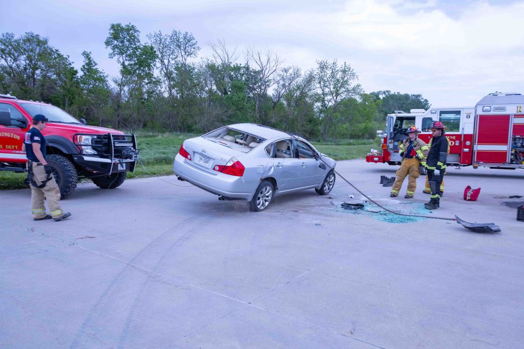 Firefighters responding to a car accident scene with a damaged silver sedan, emergency vehicles in the background.