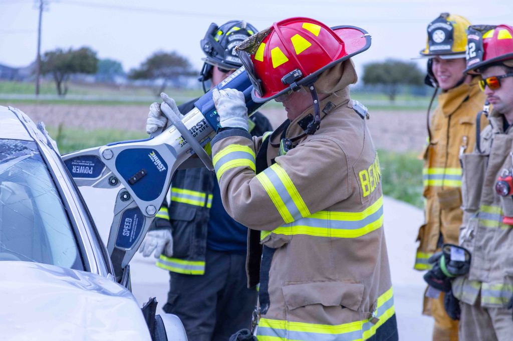 Firefighter using hydraulic rescue equipment to extricate an injured person from a vehicle, with other firefighters observing.
