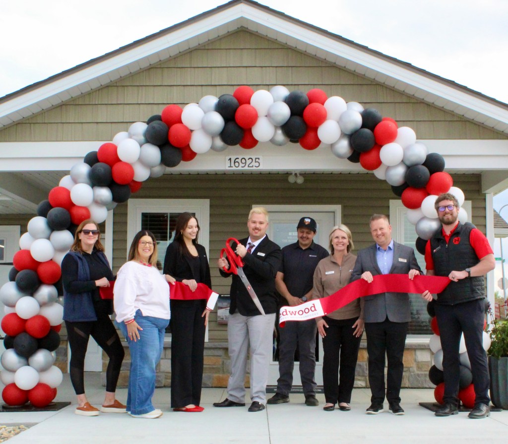 A group of seven people standing in front of a house under an arch of red, black, and white balloons, holding a large red ribbon and scissors for a ribbon-cutting ceremony.