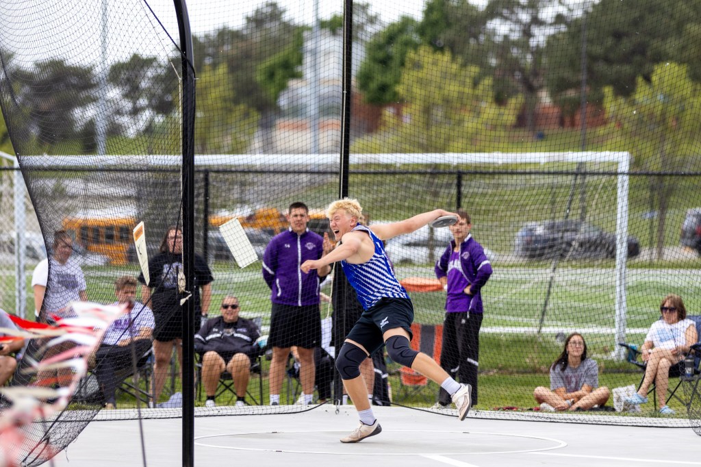 A young athlete in a blue and white striped tank top is throwing a discus during a track and field event, with spectators and coaches in the background.