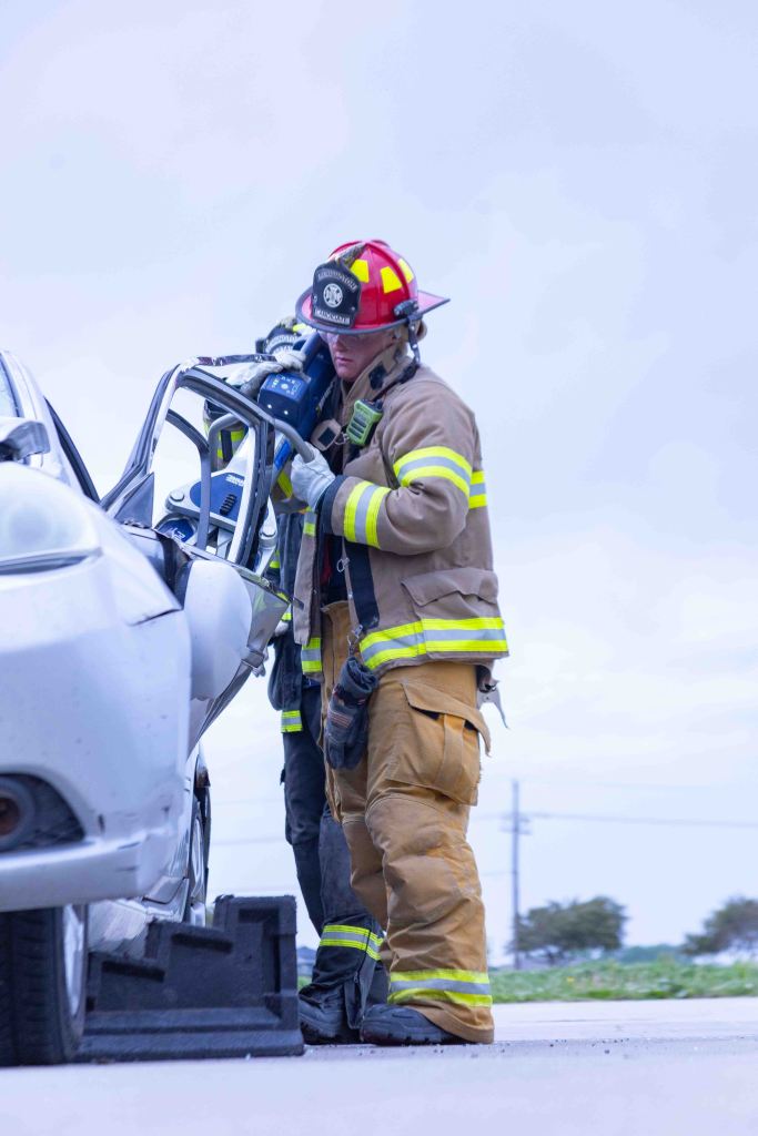 Two firefighters working on a car rescue operation, one using a hydraulic tool to remove the car door.