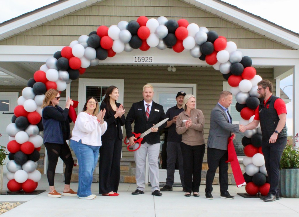 A group of people celebrating a ribbon-cutting ceremony in front of a house, with a balloon arch in red, black, and white colors. One person holds scissors ready to cut the ribbon, while others applaud.