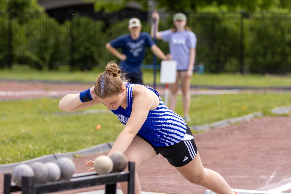 A female athlete in a blue and white sports uniform prepares to throw a shot put during practice, with two other individuals in the background observing.