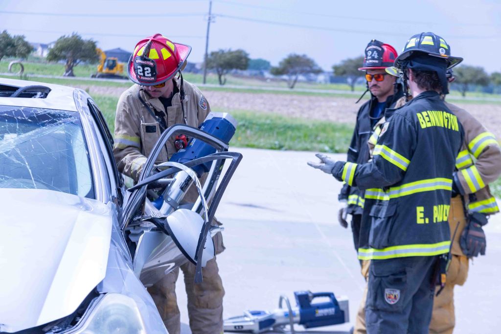 Firefighters using equipment to rescue a person from a car involved in an accident, with a damaged vehicle in the foreground.