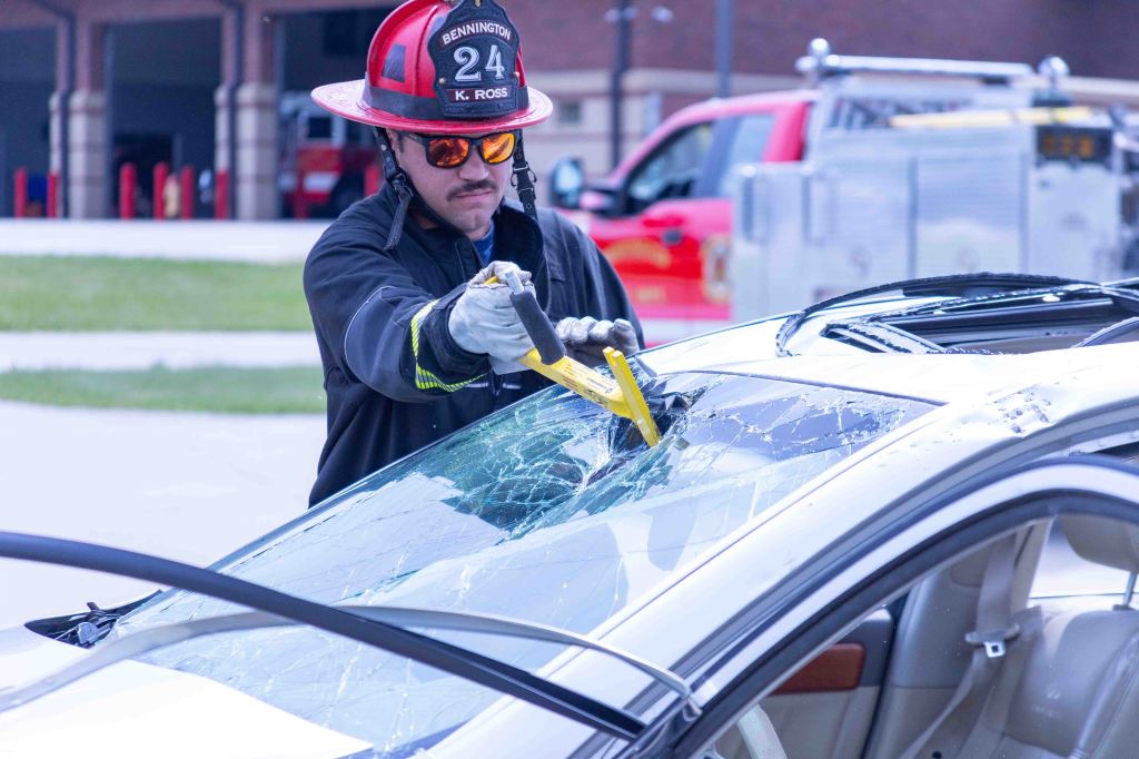 A firefighter using a tool to break through a damaged windshield of a car during a rescue demonstration.