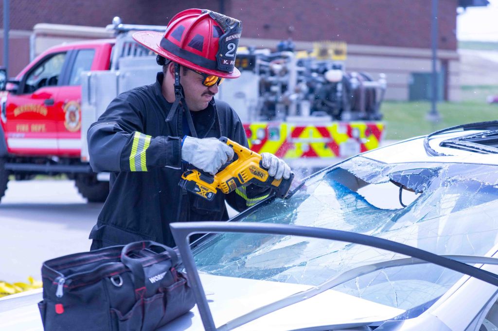 A firefighter in full gear uses a power tool to remove the windshield from a damaged car during a rescue operation, with a fire truck visible in the background.