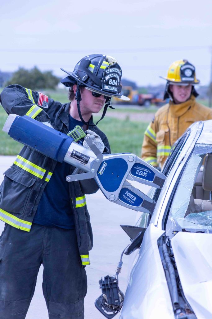 A firefighter using hydraulic tools to extricate someone from a damaged car while another firefighter observes.