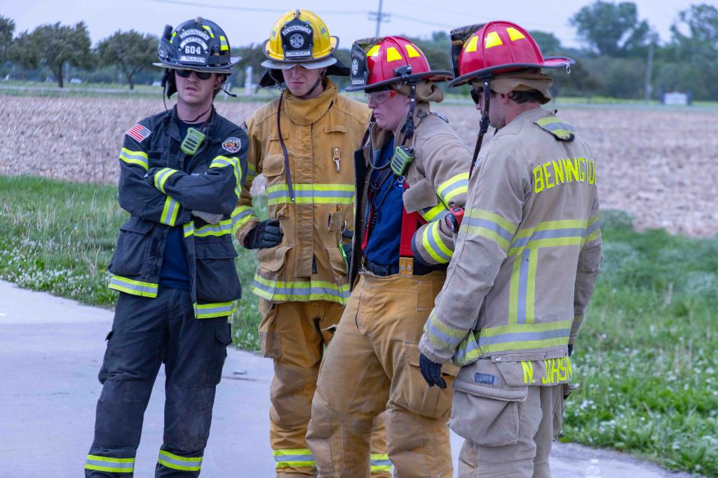 Group of four firefighters in uniforms discussing on a rural road, with fields in the background.