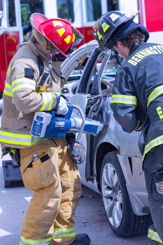 Two firefighters using hydraulic equipment to assist in a vehicle extrication, with a fire truck in the background.
