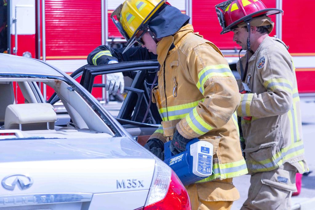 Two firefighters using tools to extricate a person from a car during a rescue operation, with a fire truck in the background.