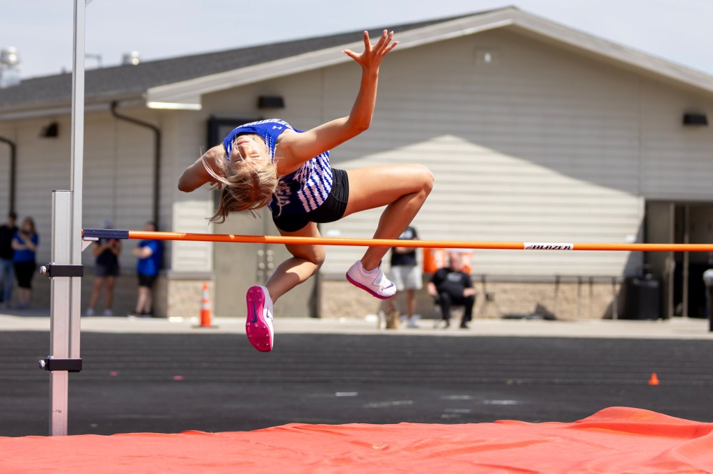 Female high jumper in mid-air over a bar during a track and field event, with a school building and spectators in the background.