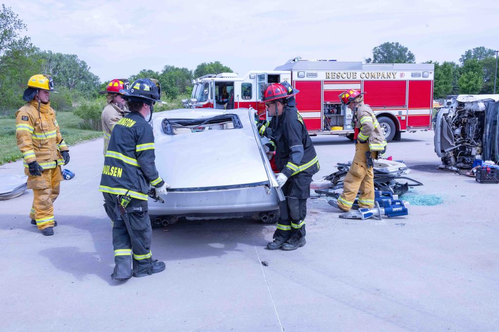 Firefighters responding to a car accident, lifting the roof off a vehicle while rescue equipment is visible in the background.