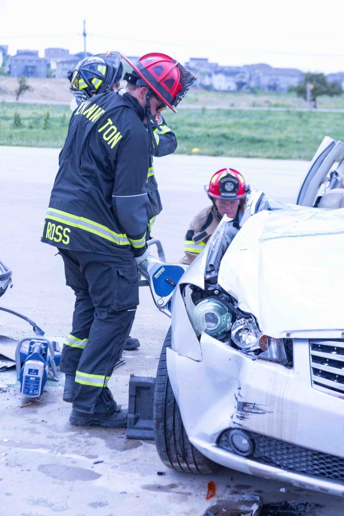 Firefighters using specialized tools to extricate an individual from a damaged white car after an accident.
