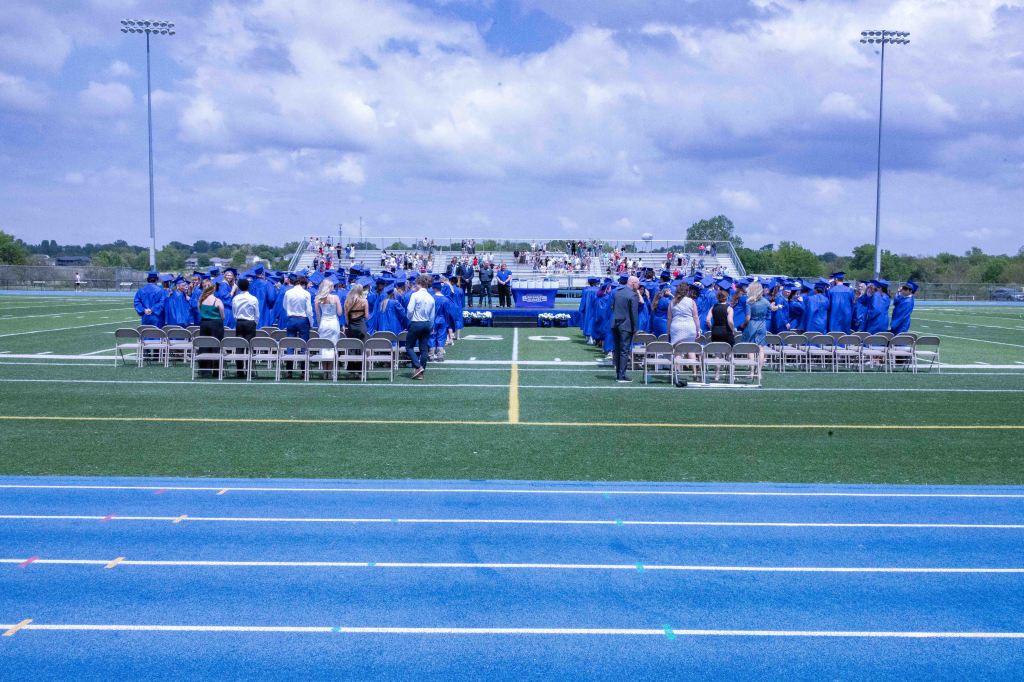 A graduation ceremony on a football field, featuring graduates in blue caps and gowns, seated guests, and a stage in the background under partly cloudy skies.