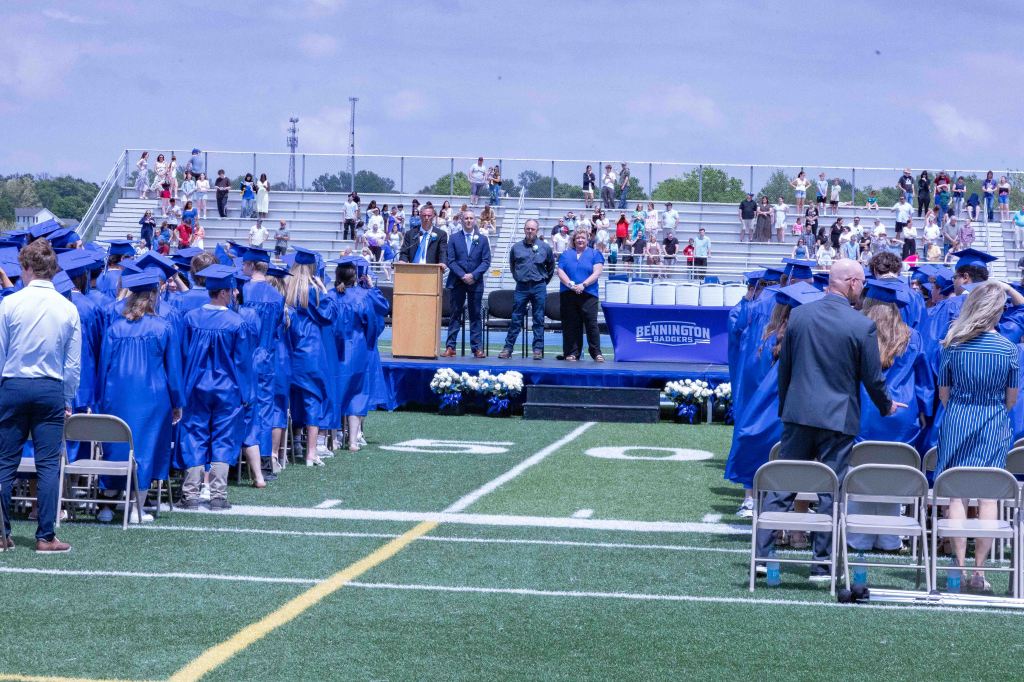 A graduation ceremony taking place on a sports field, with graduates in blue gowns and caps facing a podium. Spectators are seated in the background, and a banner for Bennington Badgers is visible.