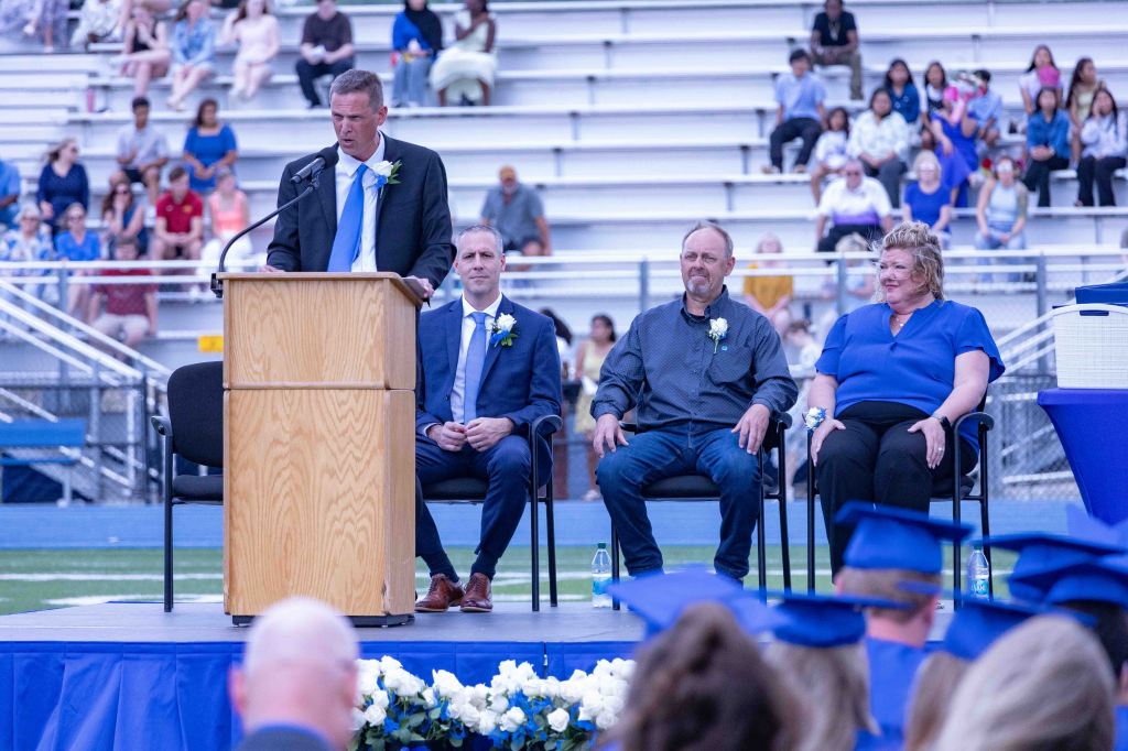 A graduation ceremony scene with a speaker at a podium and four seated individuals, including three audience members in blue caps and gowns. The background features an audience in a stadium setting.