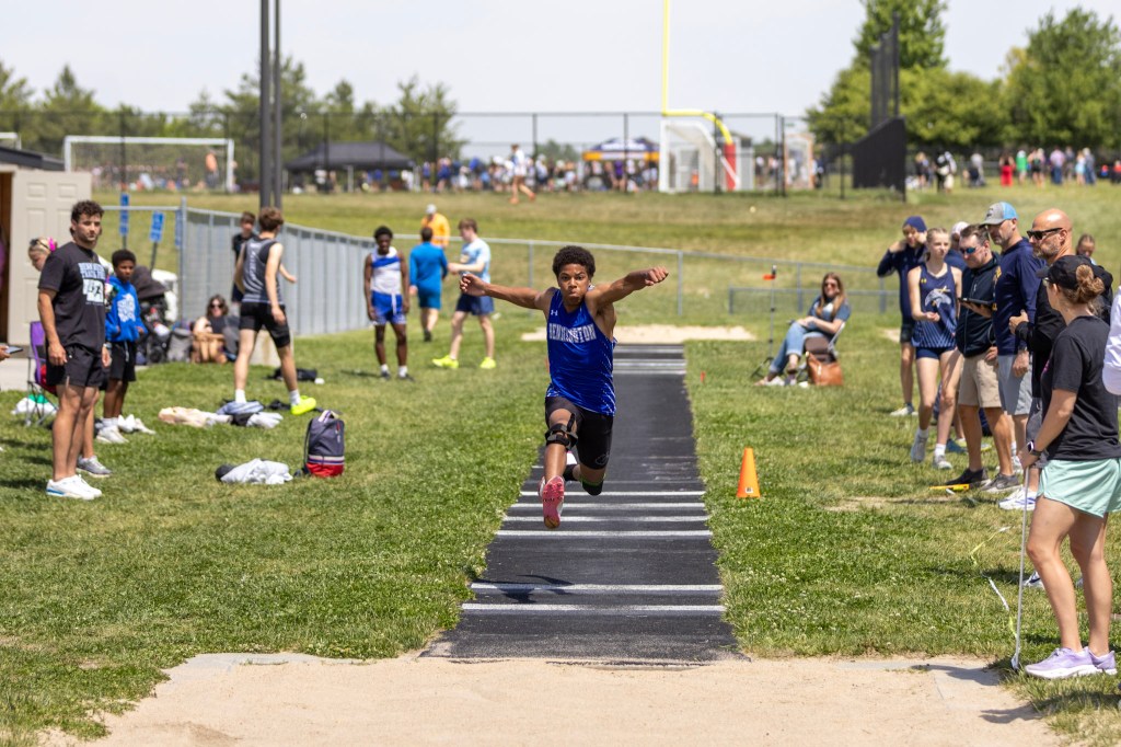 A young male athlete competing in a long jump event, wearing a blue sports uniform, leaping off a track towards a sandpit, with spectators and other athletes in the background.