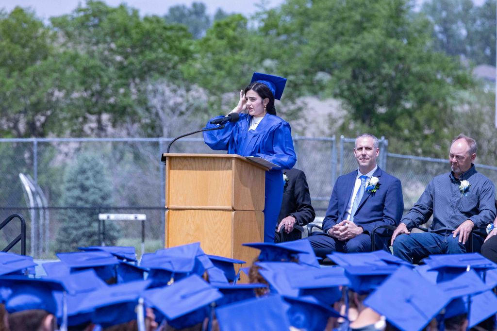 A female graduate in a blue cap and gown stands at a podium, giving a speech at a graduation ceremony, while seated attendees listen.