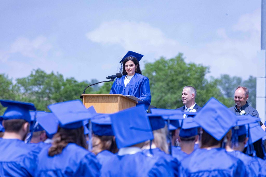 A female graduate in a blue cap and gown stands at a podium speaking to an audience of graduates, also in blue caps and gowns, under a clear sky.