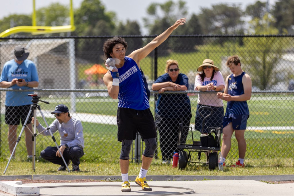A male athlete in a blue jersey is preparing to throw a shot put, with spectators watching in the background at a sports event.