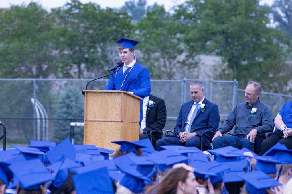 A male student in a blue graduation gown speaks at a podium during a graduation ceremony, with fellow graduates and guests seated in front of him.